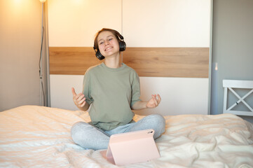 A teenage girl wearing headphones sits on her bed in casual home clothes, happily watching or listening to a tablet, illustrating digital leisure and a cozy, joyful home atmosphere