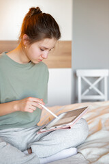 A teenage girl sits on her bed in comfortable home clothes, focused on a tablet, illustrating everyday leisure, digital technology, and a cozy home atmosphere in a relaxed setting