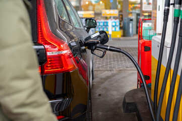 A partial view of a car with a fuel nozzle inserted into the tank at a gas station, showing the refueling process, human interaction with vehicles, and urban infrastructure development for automobiles