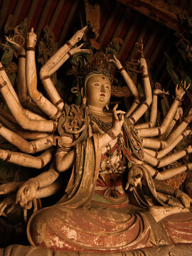 View of intricate, weathered statue of a deity with multiple arms in a dimly lit space, showcasing rich textures and spiritual depth, Jinzhong, Shanxi, China.