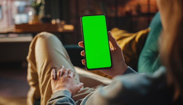 Vertical Screen: man Hand Holding on Smartphone with Green Screen Mock Up Display. Female Resting at Home and Checking Social Media on Mobile Device. Close Up Over the Shoulder Footage