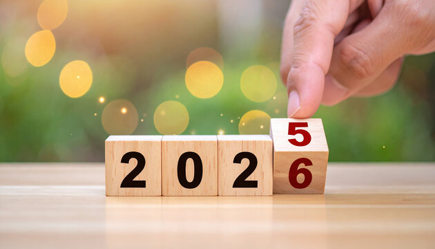 Wooden blocks displaying transition from 2025 to 2026, with hand turning block. background features bokeh effect with warm lights, creating festive and hopeful atmosphere
