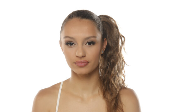 Studio shot of a young woman with brown wavy hair in a high side ponytail wearing a white strap top on a white background.