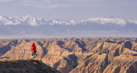 A person in a bright red jacket and a small dog stand on the edge of a cliff, offering a breathtaking view of the vast, rugged landscape of Charyn Canyon. 