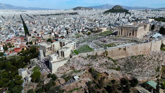 Dynamic zoom-out and lateral move over the Acropolis summit, revealing the Parthenon, tourists, Lycabettus Hill, and the vast Athens skyline.