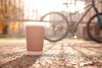 Coffee in a paper cup against the background of a bicycle on the street, lifestyle concept with sun rays and glare.