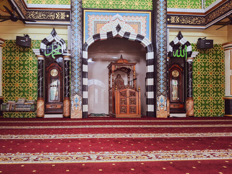 Interior detail shot of an ornate prayer niche and minbar with Islamic decoration inside of a mosque showing intricate craftsmanship, vibrant colors, and a prayer setting.