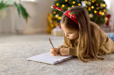 Child writing Christmas letter in notebook at home, lying on carpet by Xmas tree with magic...
