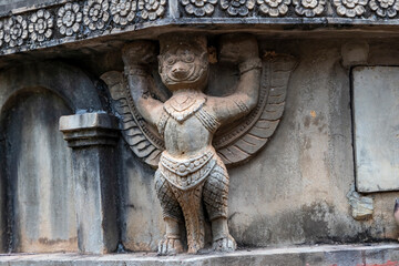 Stone sculpture of a mythical winged creature supporting a temple structure. Intricate carvings, weathered texture, solemn atmosphere. Suitable for cultural, architectural, and travel photography