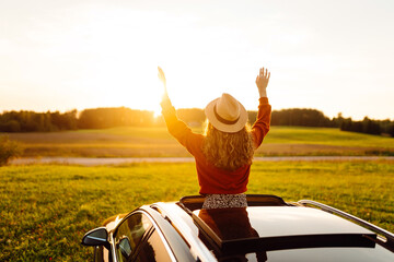Obraz premium A joyful woman leans out of a car window on a sunny day at sunset. A young woman in a hat enjoys nature and feels the freedom of a road trip. Concept of adventure and relaxation.