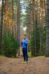 Fototapeta premium Man wearing sportswear and an orange headband standing on a pine forest path, holding and looking down at his smartphone, possibly checking a map or fitness app