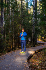 Obraz premium Man on winding wooden trail in autumn park checks his smartwatch before a run, surrounded by evergreen trees and fallen leaves, focused on fitness and training