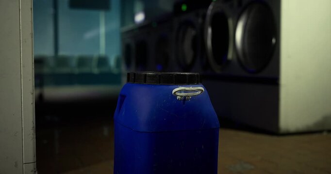 A blue container stands in the washing area, positioned in front of several laundry machines. The setting is bright with reflections, indicative of a busy laundromat.