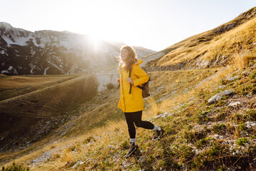 Obraz premium A joyful woman in a yellow coat and with a backpack stands on a hill and enjoys the mountain scenery. The hiker feels free and walks along a hiking trail at sunset. Nature and hiking concept.