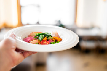 Colorful, gourmet plated pasta dish with fresh vegetables and possibly salmon, held in a hand for an appetizing food shot with selective focus and natural light.