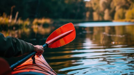 A close-up of a hand holding a red kayak paddle above calm water. Lush green trees line the riverbank in the background, reflecting in the water.