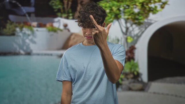 Man shows middle finger with bare finger, visible hand and uneasy grimace at a building poolside arch near water and plants; defiance.