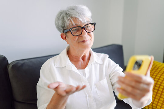 An elderly woman with gray hair and glasses waves during a video chat on her smartphone. A smiling adult blogger stands with a phone in a cozy room. Concept: atmosphere, technology, and blogging.