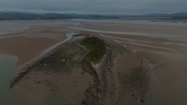 A drone flyover of a tidal sandbank island at low tide, showing surrounding mudflats and shallow water channels under an overcast sky.