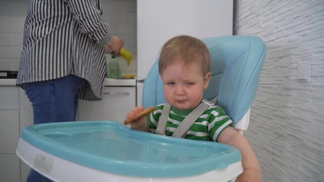 toddler boy in green striped shirt eating rusk while sitting in light blue high chair, crumbs on his face, mother standing nearby in modern kitchen