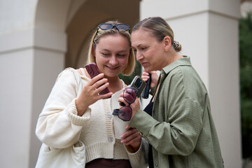 Two women smile as they look at a phone together outdoors, enjoying a light moment and sharing something on the screen.