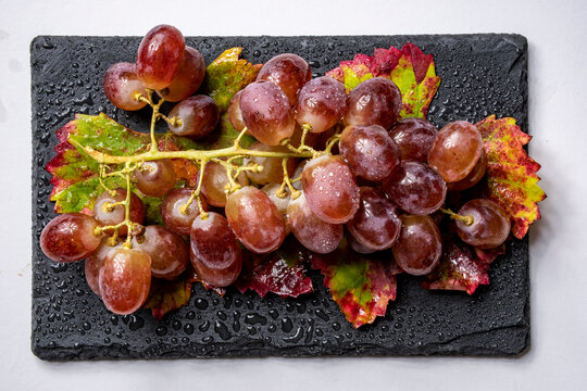 Uvas rojas frescas dispuestas en una superficie de pizarra negra, creando un castraste visual con gotas de agua en las hojas de la parra a&ntilde;aden un aspecto fresco y apetitoso