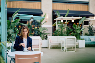 Young woman stirring coffee in a cup while sitting at an outdoor cafe table, having a break with a smartphone nearby