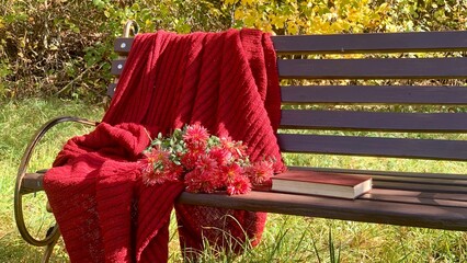 Cozy autumn scene with a red blanket, flowers, and a book on a park bench surrounded by golden leaves