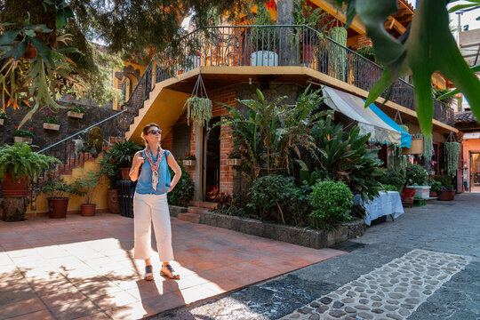 A sunlit courtyard in Tepoztlán, Mexico, surrounded by lush plants with a woman standing in the warm afternoon light looking at the rustic architecture  and tropical greenery. - Powered by Adobe