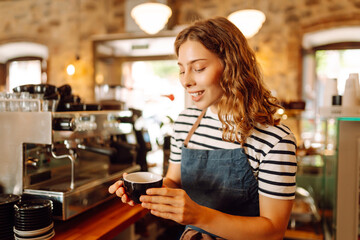 A female barista stands behind the counter with freshly brewed coffee in a cozy cafe. A smiling woman prepares and serves hot drinks inside. The concept is cozy, drinks, and small business.
