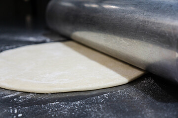 Close-up of pizza dough being rolled out with a large heavy-duty steel rolling pin on a clean stainless steel worktable.Smooth dough texture, metallic reflections, and authentic cooking process.
