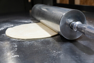 Close-up of pizza dough being rolled out with a large heavy-duty steel rolling pin on a clean stainless steel worktable.Smooth dough texture, metallic reflections, and authentic cooking process.