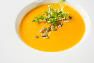 A vibrant orange pumpkin or carrot cream soup, garnished with fresh greens and scattered pumpkin seeds, served in a white bowl