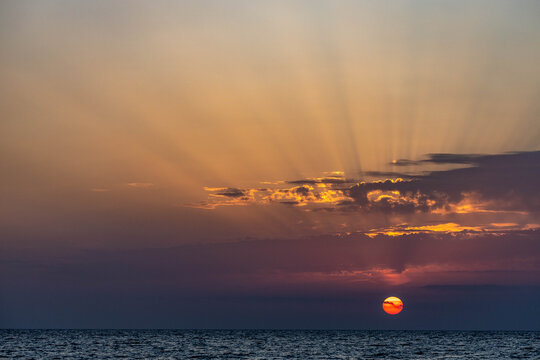 A minimalist dramatic sunset over the Mediterranean Sea, featuring a red sun disk with radial light rays through layered clouds against a gradient sky. Antalya, Turkey.


