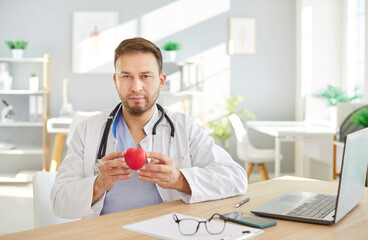 Professional focused male cardiologist wearing stethoscope, showing red heart toy to camera,...