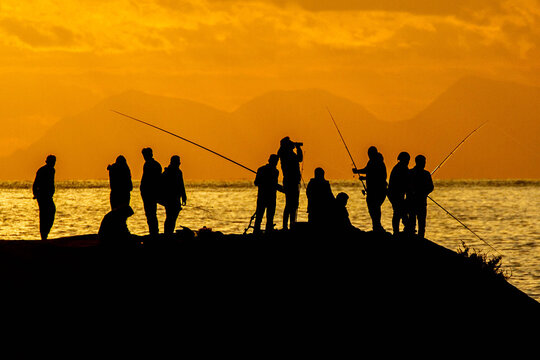 Silhouetted fishermen on a rocky breakwater at sunset, casting lines into the orange Mediterranean waters. A traditional fishing scene in Evrenseki, Antalya, Turkey.

