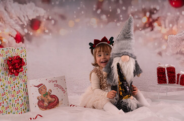 Kleines zweij&auml;hriges M&auml;dchen tr&auml;gt ein Kleid mit goldenen Pailletten und hat einen Weihnachtswichtel in der Hand.