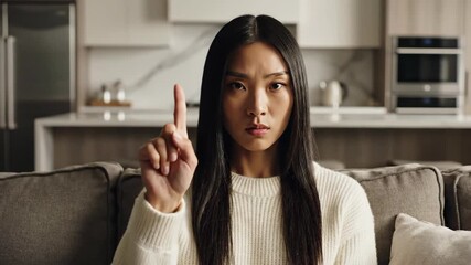 Serious Asian woman raising index finger in warning while explaining concept in modern living room, ideal for online courses and lifestyle content