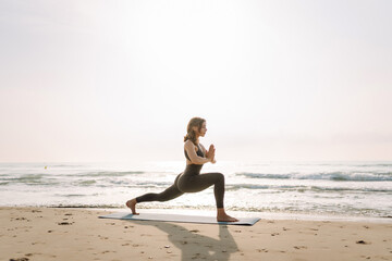 A young woman in athletic clothing practices yoga on a blue mat on the beach. The beautiful woman practices various exercises and enjoys the seascape. Concepts of yoga, relaxation, and meditation.