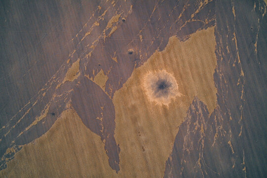 Aerial drone shot showing the massive impact crater and blast zone left by an explosion in a field. The abstract pattern illustrates war destruction, bomb damage, and the tragedy of conflict.