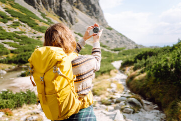 Rear view of female hiker, holding backpack and compass, on rocky trail in background. Hiker looks...