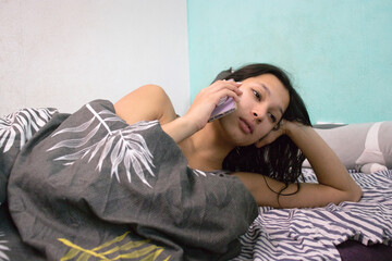 A woman lies on her side in bed, propping her head with one hand and holding a smartphone in the other, smiling softly. She is wrapped under a gray blanket, looking relaxed and content.