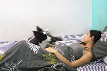 A young woman is lying in bed under a patterned blanket, interacting with a black and white Border Collie dog. She looks relaxed and happy, reaching out towards the playful dog beside her.