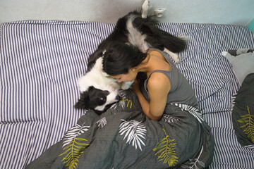 A woman lays sideways on her bed, smiling, while her playful Border Collie dog stands over her, creating a lively and affectionate atmosphere in the bedroom.