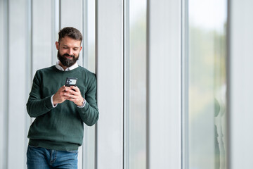 Fototapeta premium Man smiling using phone leaning on modern window wall
