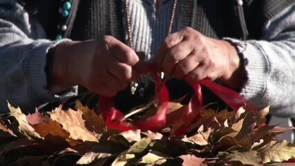 Close-up of hands crafting a wreath with autumn leaves and red ribbon, a festive fall decoration.