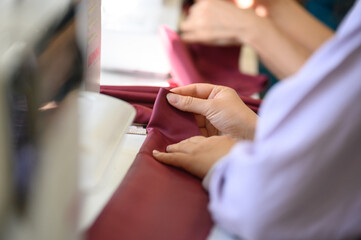 A seamstress's hands work on burgundy fabric on a sewing machine in a studio.