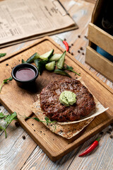 A savory grilled meat patty sits on a wooden plate, topped with a dollop of green sauce. Fresh cucumbers and a small bowl of dark sauce accompany the dish, highlighting its flavors