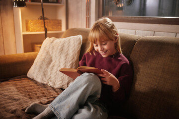 Obraz premium A child sits on a comfortable couch, deeply engaged with a book. The warm lighting of the room adds to a cozy atmosphere during the evening, with soft textures surrounding her.