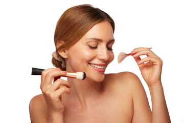 A cheerful woman applies makeup with a brush, radiating confidence and joy. Her clear skin and bright smile highlight her skincare and beauty care routine in a well-lit environment.